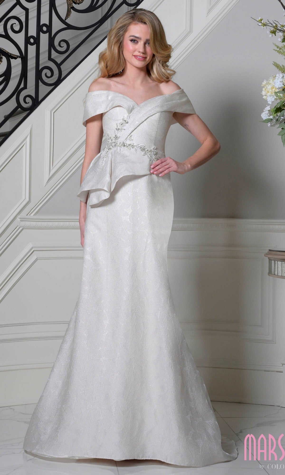 A woman in a champagne Marsoni MV1397 long formal dress with floral embroidery and an asymmetrical peplum waist stands indoors near a staircase.